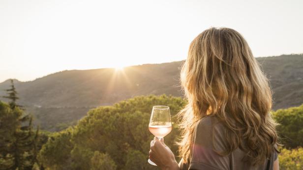 Eine Frau hält ein Glas Wein und beobachtet den Sonnenuntergang über den Hügeln bei Portoferraio, Toskana, Italien.