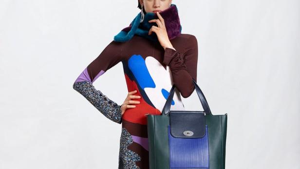 Eine Frau in einem langen, bunten Kleid mit einer großen Handtasche posiert vor einem weißen Hintergrund.