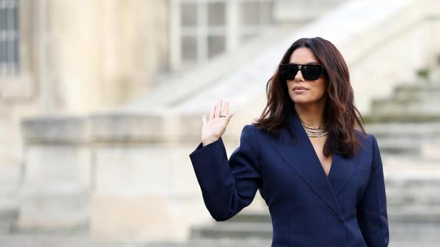 Eine Frau mit Sonnenbrille, Blazer und Leggings posiert auf einem gepflasterten Platz.