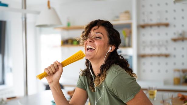 Eine Frau singt mit einem Bund Spaghetti in der Hand in ihrer Küche.
