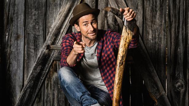 Ein Mann mit Hut und kariertem Hemd lehnt an einer alten Axt und zeigt auf den Betrachter.