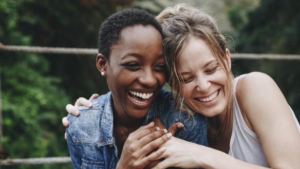 Zwei lachende Frauen umarmen sich vor einem grünen Hintergrund.