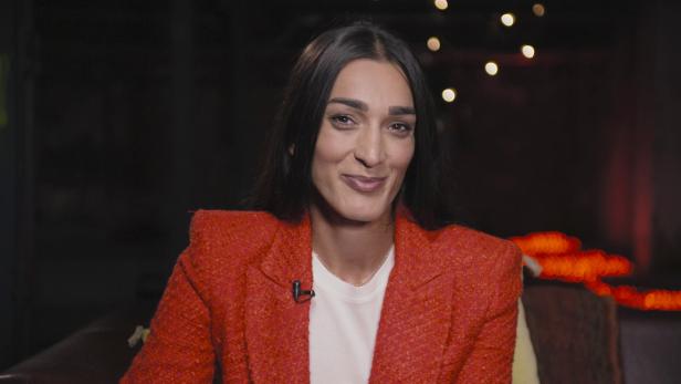 Eine Frau mit dunklen Haaren trägt ein rotes Jackett und lächelt.