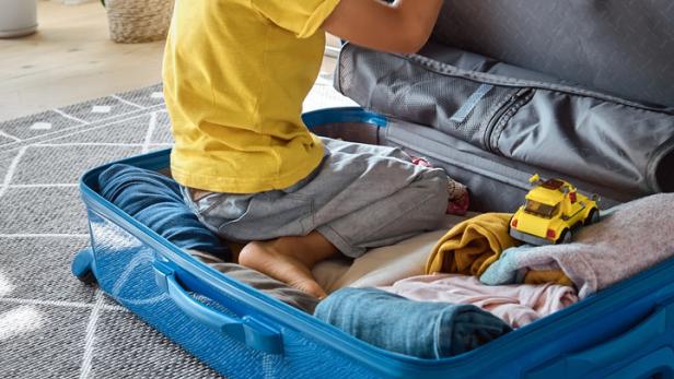 Ein Junge sitzt in einem blauen Koffer, umgeben von Kleidung und einem Spielzeugauto.