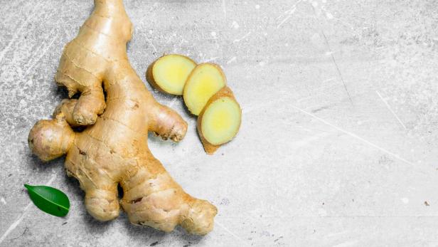 Eine Ingwerwurzel liegt neben drei Ingwerscheiben und einem grünen Blatt auf einer grauen Oberfläche.