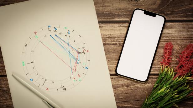 Ein Horoskop, ein Stift, ein Smartphone und Blumen liegen auf einem Holztisch.