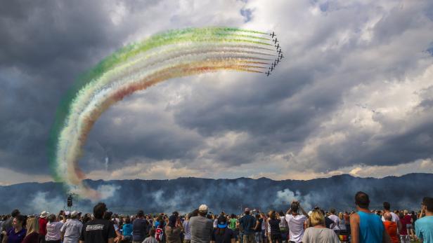 Eine Gruppe von Flugzeugen zieht farbige Rauchspuren vor einem bewölkten Himmel, während Zuschauer zusehen.