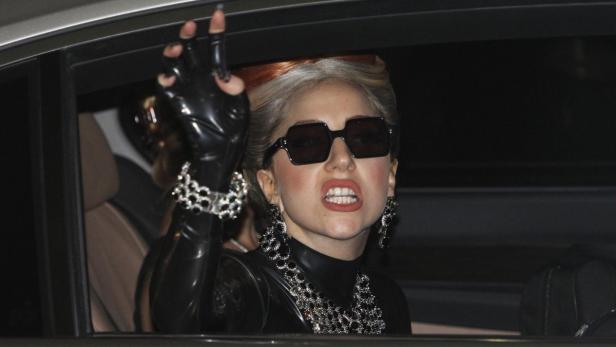 Lady Gaga in einem schwarzen Outfit und Sonnenbrille gestikuliert aus einem Auto.