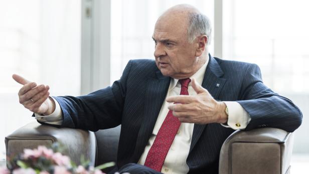 Ein älterer Mann im Anzug gestikuliert, während er in einem Sessel sitzt.