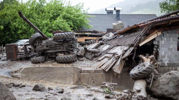 Ein umgestürzter Traktor und Trümmer vor einem beschädigten Haus nach einer Überschwemmung.