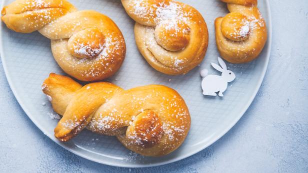 Ein Teller mit süßen, hasenförmigen Brötchen, bestäubt mit Puderzucker, neben einem Nest mit bunten Ostereiern.