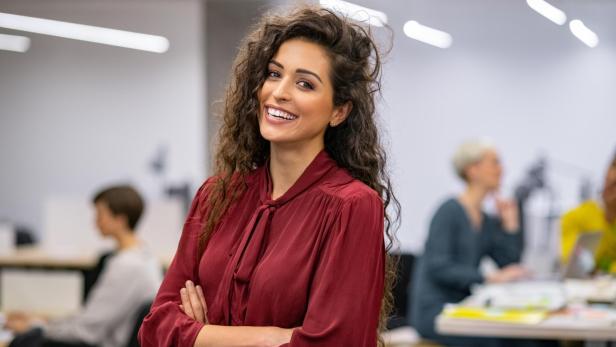 Eine lächelnde Frau mit lockigem Haar und roter Bluse steht in einem Büro.