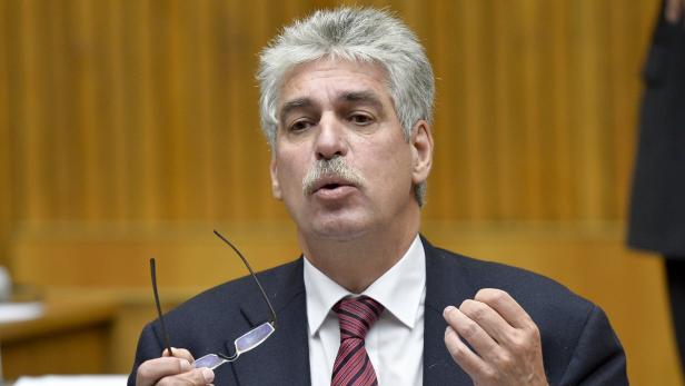 Ein Mann mit grauem Haar und Schnurrbart hält eine Brille in der Hand und gestikuliert.