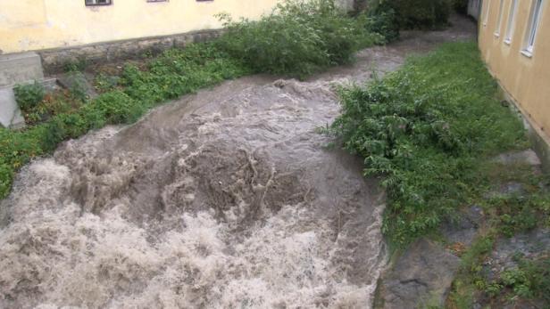 Ein reißender, schlammiger Fluss fließt zwischen zwei Gebäuden hindurch.