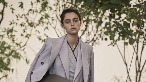 Eine Frau in einem hellgrauen Hosenanzug mit einer übergroßen Tasche steht auf einer Holzterrasse.