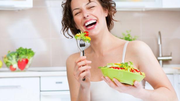 Eine lachende Frau isst einen Salat in einer hellen Küche.