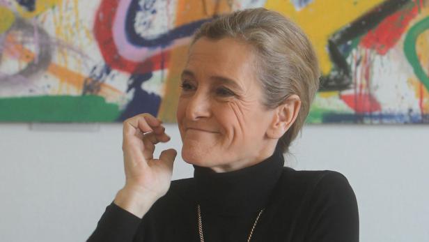 Eine Frau mit schwarzem Rollkragenpullover und Goldkette vor einem bunten Gemälde.