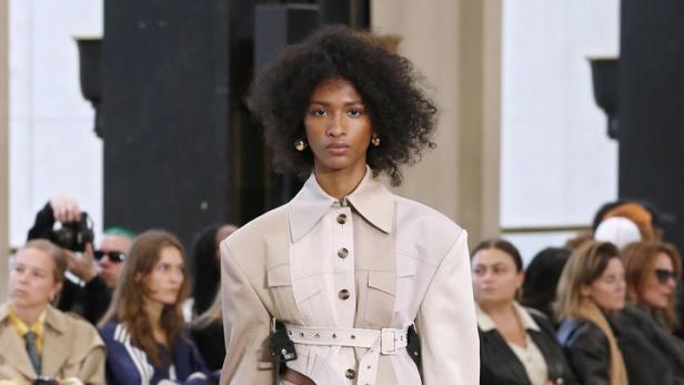 Eine Frau präsentiert ein beigefarbenes Outfit mit Plisseerock auf einem Laufsteg.