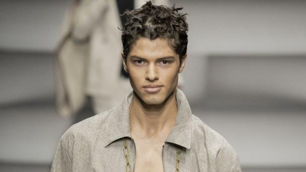 Ein Mann mit offenem Hemd und einer Handytasche an einer goldenen Kette auf einem Laufsteg.