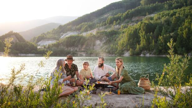 Eine Familie grillt am Ufer eines Sees vor einer bewaldeten Hügellandschaft.