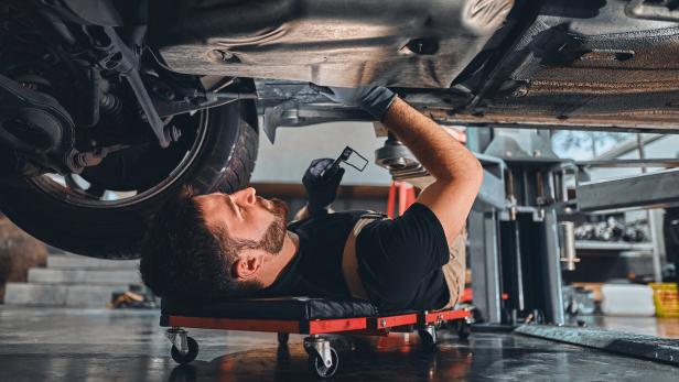 Male car mechanic worker working using wrench tool for repair, maintenance underneath car. Mechanic vehicle service checking under car in garage.