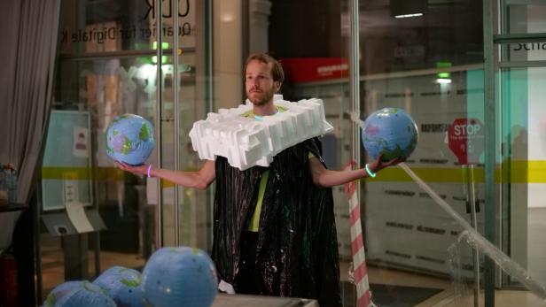 Ein Mann in einem Kostüm aus Müll und Styropor hält zwei Globen in den Händen.