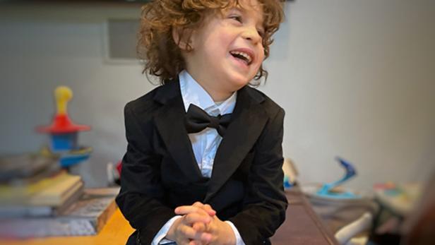 Ein lachender Junge mit lockigem Haar, gekleidet in Anzug und Fliege, sitzt auf einer Truhe.