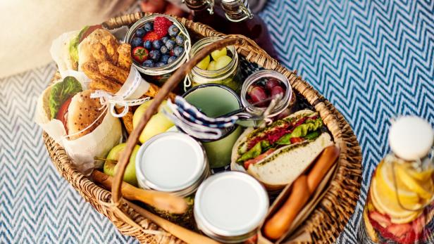 Ein Picknickkorb gefüllt mit Sandwiches, Obst und Getränken auf einer blau-weiß gemusterten Decke.
