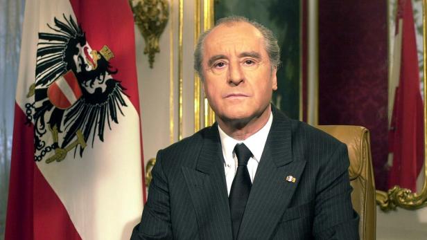 Ein Mann im Anzug sitzt an einem Tisch vor einer österreichischen Flagge mit Bundesadler.