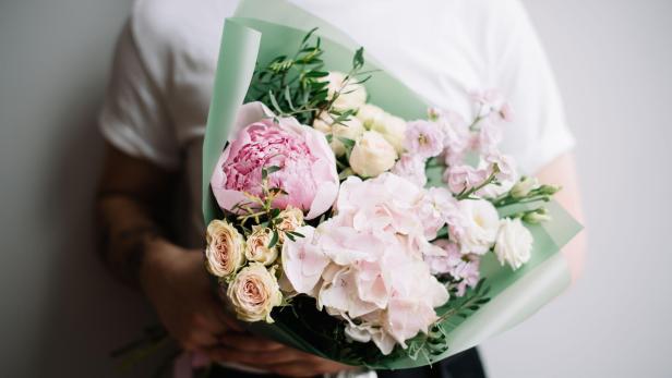 Very nice young man in a white t-shirt holding a beautiful blossoming flower bouquet of fresh peony, hydrangea, roses, mattiola in tender pink colours on the grey wall background