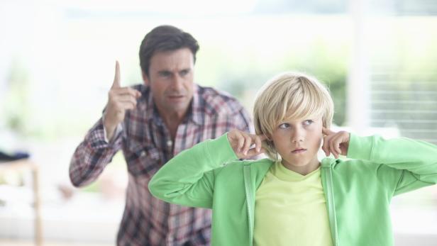 Ein Junge hält sich die Ohren zu, während ein Mann im Hintergrund mit erhobenem Zeigefinger schimpft.