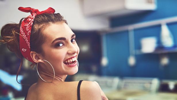 Eine lächelnde Frau mit rotem Haarband und großen Ohrringen blickt über ihre Schulter.