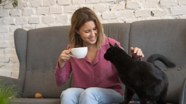 Eine Frau sitzt auf einem Sofa, hält eine Tasse und streichelt eine schwarze Katze.
