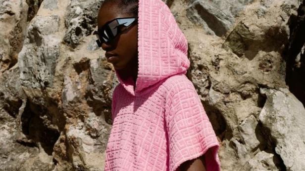 Eine Frau mit Sonnenbrille und einem rosa Givenchy-Strandponcho mit passender Tasche vor einem Felsen.