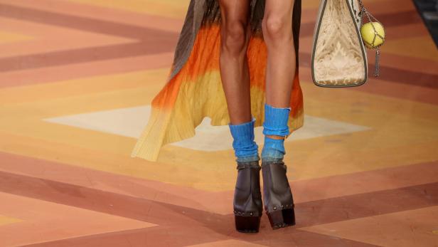 Nahaufnahme von Beinen mit blauen Socken und Plateauschuhen, dazu eine Handtasche mit Apfelanhänger.