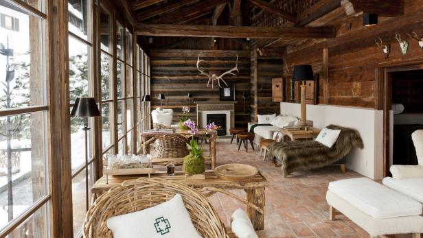 Ein rustikales Wohnzimmer mit Holzwänden, Kamin und großen Fenstern mit Blick auf eine verschneite Landschaft.