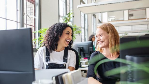 Zwei Frauen sitzen lachend an ihren Schreibtischen in einem Büro.