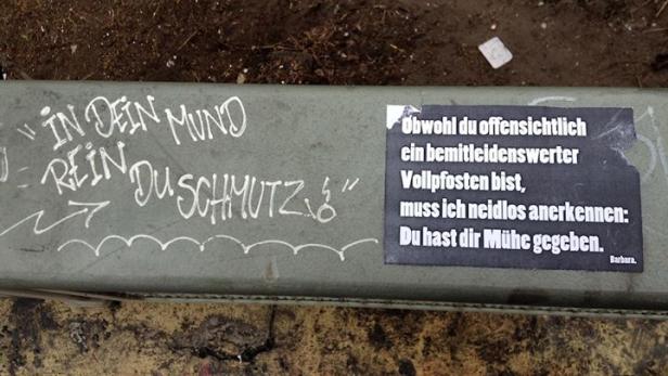 Auf einer Bank befinden sich Graffiti und ein Aufkleber mit dem Text „Obwohl du offensichtlich ein bemitleidenswerter Vollpfosten bist…“.