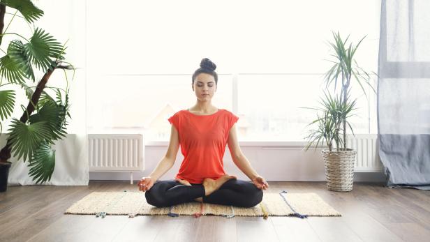 Eine Frau meditiert in einem hellen Raum im Lotussitz auf einer Matte.