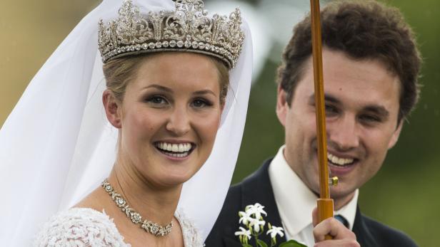 Eine strahlende Braut mit Diadem und Schleier, neben ihr ein lächelnder Mann mit Schirm.