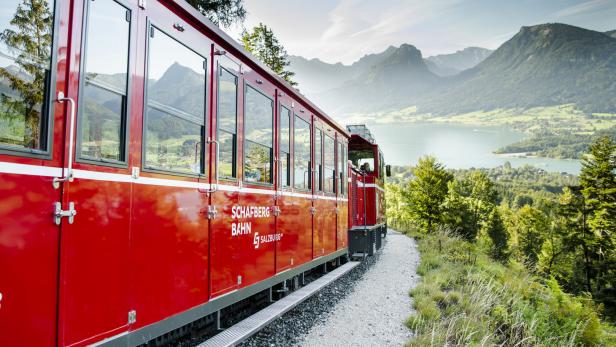 Die rote Schafbergbahn fährt vor einer Bergkulisse und einem See entlang.