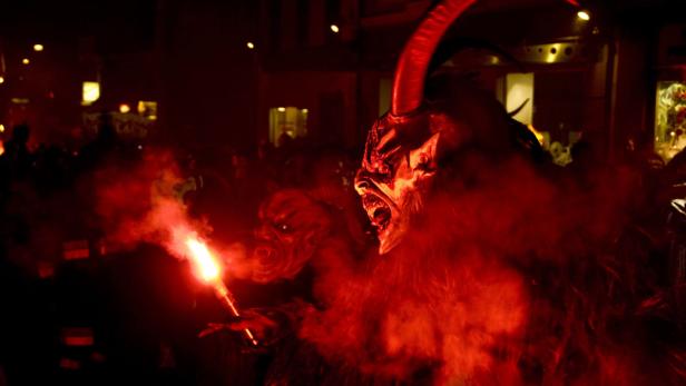 Krampusgestalten mit Hörnern und Fell stehen im roten Rauch, einer hält eine Fackel.