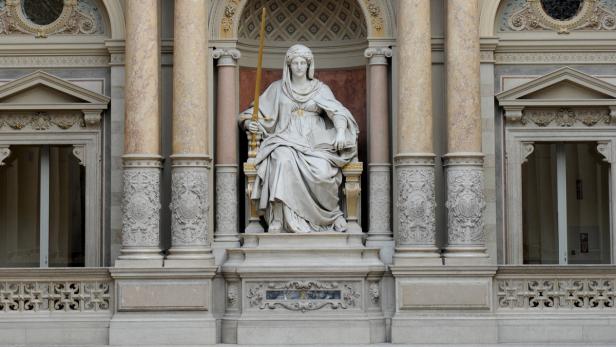 Eine Statue der Justitia mit Schwert und Buch, sitzend vor einem Gebäude mit Säulen.