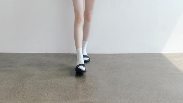 Eine junge Frau mit langem Haar trägt ein weißes Hemd, Socken und Sandalen.