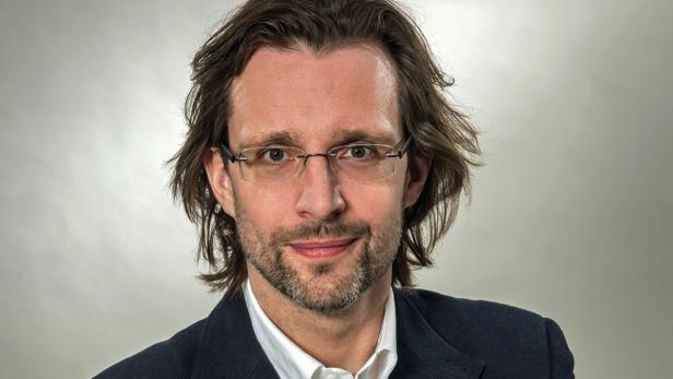 Ein Mann mit Brille und dunklem Anzug vor einem grauen Hintergrund.