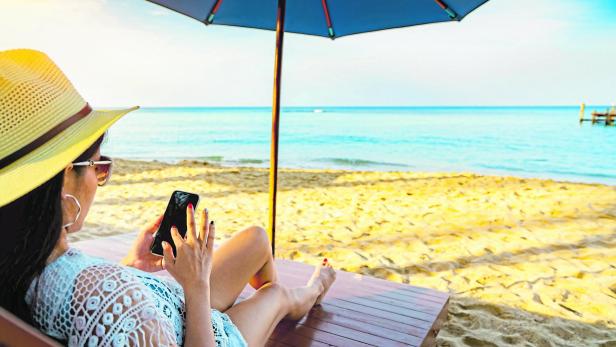 Asian woman with hat sit on sunbed under beach umbrella at sand beach and using smartphone on summer vacation. Girl in casual style relax and enjoy holiday at tropical paradise beach. Summer vibes.