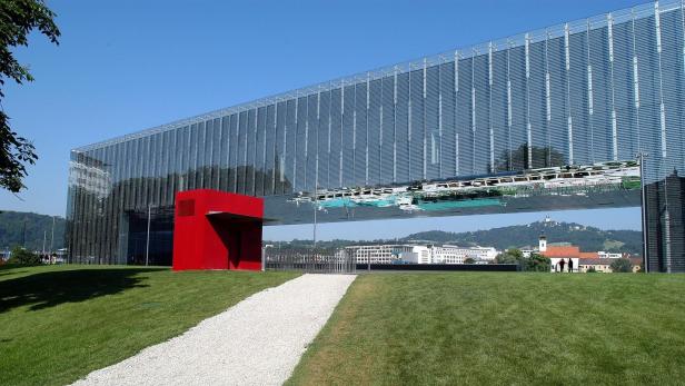Ein moderner, verglaster Bau mit spiegelnder Fassade und einem kleinen roten Kubus steht auf einer grünen Wiese.