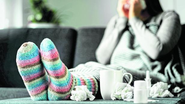 Sick woman with flu, cold, fever and cough sitting on couch at home. Ill person blowing nose and sneezing with tissue and handkerchief. Woolen socks and medicine. Infection in winter.