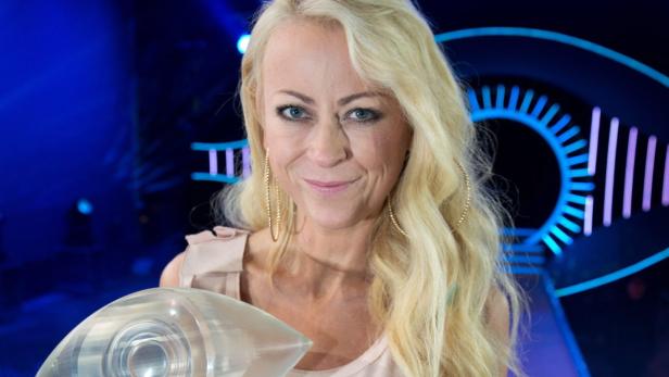 Eine blonde Frau hält eine Trophäe vor dem Hintergrund des „Big Brother“-Logos.
