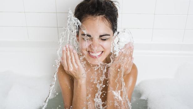 Eine Frau sitzt lächelnd in einer Badewanne und spritzt sich Wasser ins Gesicht.